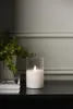 A white LED candle in a glass holder sits on a black surface next to a potted plant. Podgląd zdjęcia produktu