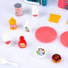 A collection of toy kitchen accessories, including various food containers, plates, and a spoon, arranged on a white surface. Produktbild-Vorschau