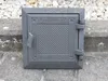 A square cast iron revision door with a textured surface and hinges, placed on a concrete surface with gravel in the background. Produktbild-Vorschau