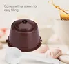 A reusable coffee capsule and a spoon are shown in the foreground, with a coffee cup with splashing coffee in the background. Produktbild-Vorschau