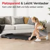 A woman is interacting with a foldable treadmill placed on a wooden floor in front of a sofa. Produktbild-Vorschau