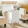 A white nightstand with three drawers is placed between two armchairs in a living room setting. A tablet and glasses are on top of the nightstand. Podgląd zdjęcia produktu
