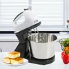 A white and black electric stand mixer with a stainless steel bowl is shown on a white countertop. Three square pieces of cake are on a white plate in the foreground. In the background, there are blinds and a glimpse of a kitchen. Produktbild-Vorschau