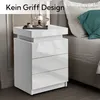 A white high-gloss nightstand with three drawers and a top storage compartment, placed next to a bed with a teal pillow. Produktbild-Vorschau