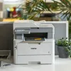 A Brother printer is shown on a desk with a plant in the background. Produktbild-Vorschau