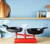 A kitchen scene with a scale holding two frying pans, a shelf with kitchenware in the background. Produktbild-Vorschau