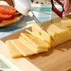 Weißes elektrisches Messer schneidet einen Block gelben Käse auf einem Holzschneidebrett, mit Tomatenscheiben im Hintergrund Anteprima dell’immagine prodotto