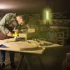 A man is working in a workshop with a power tool, illuminated by a hanging work light. Voir les images du produit
