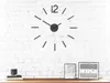 A modern, minimalist wall clock with black hour markers and hands is mounted on a white brick wall. Below the clock, on a wooden shelf, are two framed pictures and a stack of books with a small black cylinder on top. Produktbild-Vorschau