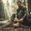 A man is tying his hiking boots in a forest setting next to a stream. Produktbild-Vorschau