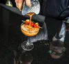 A bartender is pouring a drink into a cocktail glass, which is garnished with cherry tomatoes and cucumber. Náhled obrázku produktu