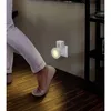 A person's legs and feet are shown walking past a wall-mounted motion-sensing night light. Produktbild-Vorschau