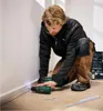 A man is kneeling on the floor, using a cordless drill to attach a white baseboard to a wall. Produktbild-Vorschau