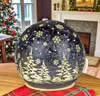 A decorative LED sphere with a Christmas theme is placed on a table in a room with a Christmas tree and fireplace in the background. Produktbild-Vorschau