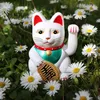 A white Maneki-neko cat figurine sits in a field of daisies. Produktbild-Vorschau