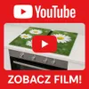 A YouTube promotional image showing a glass cutting board with a daisy pattern on a stovetop, with a play button overlay. Produktbild-Vorschau