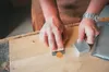 A person's hands are shown working on a wooden surface with a hand plane and a metal block. Anteprima dell’immagine prodotto