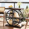 A narrow side table with a circular shelf unit is placed on a balcony overlooking a body of water and trees. The table is decorated with a bowl of fruit, a vase of flowers, books, and decorative objects. Anteprima dell’immagine prodotto