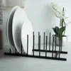 A black metal dish drying rack holding two white plates on a light-colored surface, with a white orchid in a metallic pot in the background. Produktbild-Vorschau