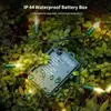 A green battery box for LED string lights is nestled among green leaves, with some lights visible. Produktbild-Vorschau