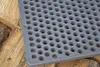 A close-up of a cast iron grate with circular holes, resting on a pile of wood. Produktbild-Vorschau