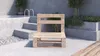 A wooden pallet bench sits on a concrete patio next to a set of concrete stairs and planters. Produktbild-Vorschau