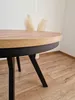 A close-up shot of a round dining table with a wooden top and black metal legs, set against a white wall and wooden floor. Produktbild-Vorschau