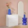 A white dresser with blue vases and flowers on top, next to an arched mirror, in a room with pink walls. Anteprima dell’immagine prodotto