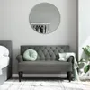 A dark gray tufted bench with green cushions and a folded green throw is placed in front of a wall with a round mirror and a bed. A plant is visible to the right. Slippers are on the rug. Produktbild-Vorschau