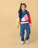 A young girl wearing a colorful sweater and jeans, with a backpack on, stands against a plain background. Produktbild-Vorschau