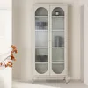 A beige display cabinet with fluted glass doors and arched tops, standing in a minimalist room with a branch of dried flowers to the left. Voir les images du produit