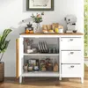 A white and wood buffet cabinet with its doors open, displaying kitchenware and food items inside. The top surface has a coffee grinder, oranges, a vase of flowers, coffee bags, and a coffee maker. Podgląd zdjęcia produktu