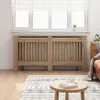 A wooden radiator cover is placed in front of a window with blinds, with a rug and a coffee table in the foreground. Voir les images du produit