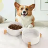 A corgi dog is lying down behind two white ceramic pet bowls, one filled with kibble and the other with water. The bowls are elevated on wooden stands. Produktbild-Vorschau
