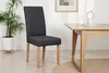 A dark gray upholstered dining chair with natural wood legs is shown next to a light wood dining table. A potted plant is visible on the left, and a framed picture hangs on the wall in the background. Produktbild-Vorschau