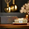A WoodWick candle and its packaging are displayed on a wooden table with decorative golden objects in the background. Podgląd zdjęcia produktu