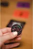 A hand holding a round pin with the word "HITSTER" on it, with colorful neon-like rings around the text. In the background, there are blurred out colorful cards and a phone. Produktbild-Vorschau