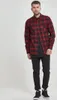 A man is standing and posing in front of a white background, wearing a red and black checkered flannel shirt over a dark t-shirt and black jeans. Produktbild-Vorschau