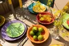 A table setting with colorful plates, cutlery, corn on the cob, limes, and yellow flowers in a vase. Náhled obrázku produktu