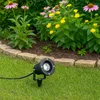A black LED garden spotlight is placed in the grass in front of a flower bed with pink and yellow flowers. Produktbild-Vorschau