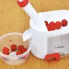 A cherry pitter is shown on a wooden surface, with cherries in the pitter and a bowl of pitted cherries next to it. Produktbild-Vorschau