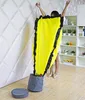 A person is holding up a large yellow toy storage bag with black trim, which is partially filled and inserted into a gray storage bin. A round gray mat is on the floor next to the bin. Shelving units with decorative items are visible in the background. Podgląd zdjęcia produktu