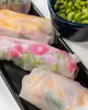 A close-up of several translucent spring rolls filled with colorful ingredients, arranged on a black rectangular tray. A small blue bowl filled with green edamame beans is visible in the upper right corner. Produktbild-Vorschau