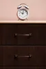 A pink alarm clock sits on top of a dark wood dresser with gold handles. Produktbild-Vorschau