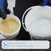 A person wearing blue gloves is holding a container of yellow liquid next to a bucket of white powder. Anteprima dell’immagine prodotto