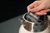 A hand is holding a black filter over an open stainless steel kettle. Podgląd zdjęcia produktu