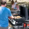 A man is grilling hot dogs and buns on a portable grill in an outdoor setting, with children playing in the background. Produktbild-Vorschau
