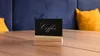 A small black sign with the word 'Coffee' written in white script sits on a wooden table. Behind the sign, a plush dark blue sofa and a yellow pillow are visible. Anteprima dell’immagine prodotto