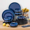 A set of blue and black stoneware dishes is displayed on a wooden surface, with lemons and a vase of yellow flowers in the background. Voir les images du produit