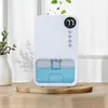 A white electric dehumidifier with a blue water reservoir sits on a wooden surface, with a plant in a white vase to the left and a blurred background. Produktbild-Vorschau